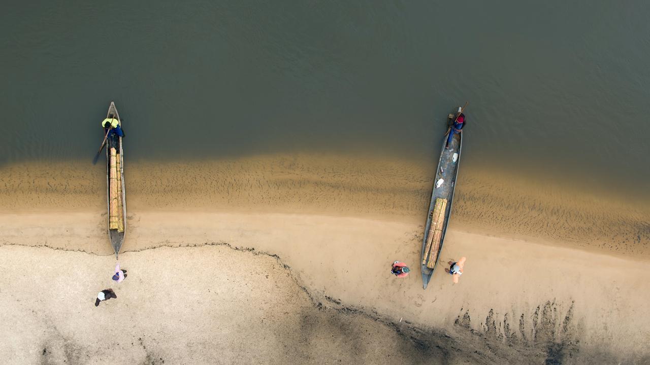 Zwei mit Papyrus-Bündeln beladene Boote auf einer Sandband, sie liegen zur Hälfte im Wasser. Vier Männer befinden sich am Ufer, jeweils einer im Bug der Boote.