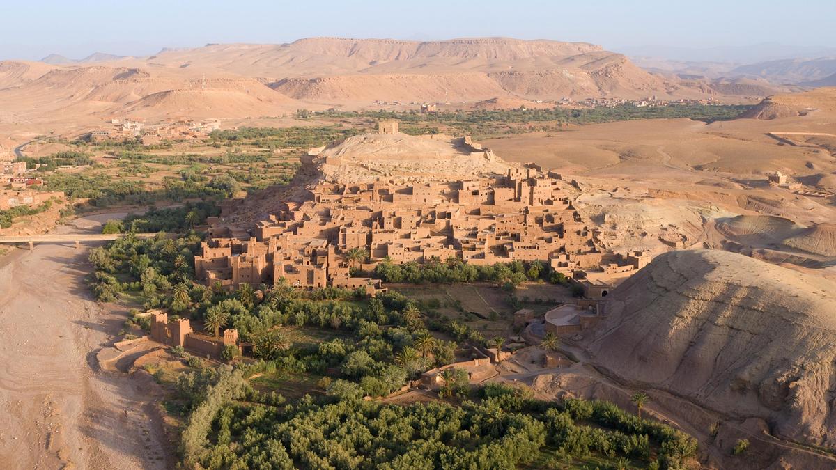 Die rötlichen Lehmmauern einer befestigten Stadt ruhen auf Felsen. In der Umgebung befinden sich einige rötliche Berge. Bepflanzte grüne Flächen bilden einen Kontrast zum roten Sand und den Felsen.