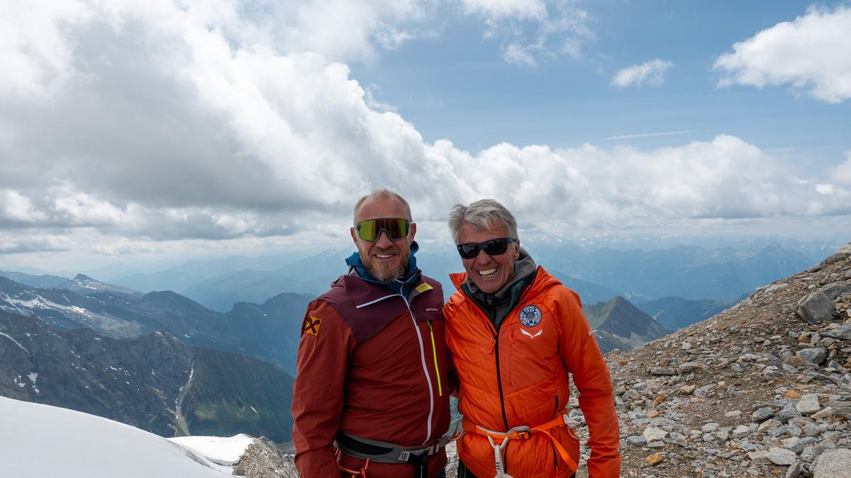 Hermann Maier mit weinroter Jacke, Peter Habeler trägt orange. Beide haben Sonnenbrillen auf und lachen in die Kamera, im Hintergrund eine Geröllhalde und hohe Berge. Große Wolken bedecken den blauen Himmel fast vollständig.