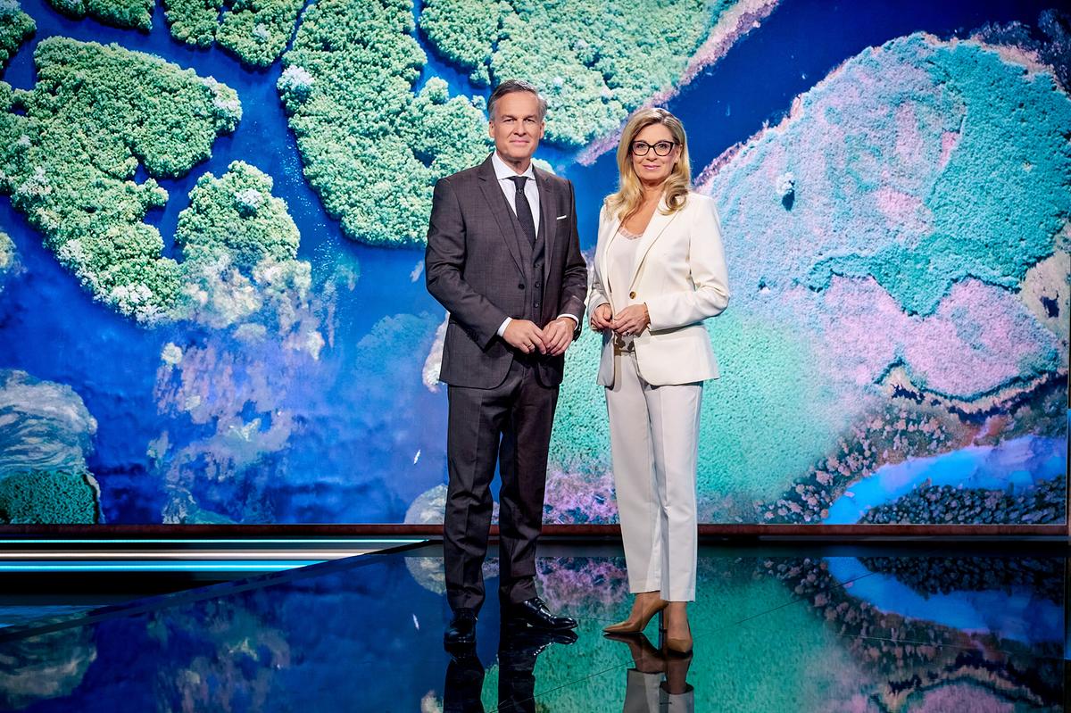 Christa Kummer im hellen Anzug und Tarek Leitner im dunklen Anzug vor einer Videowand, auf der Gewässer und Inseln von oben dargestellt sind.