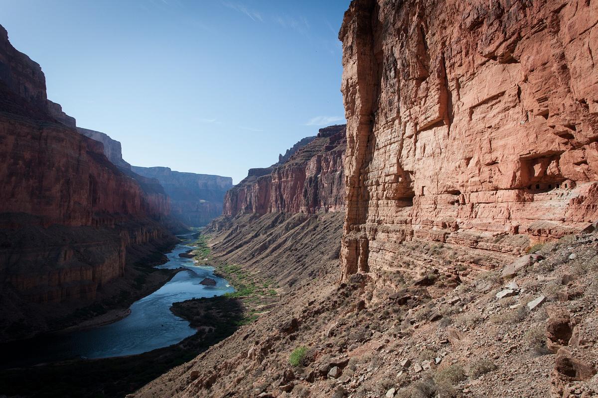 In den steilen, roten Felswänden des Canyons sind Höhlenöffnungen erkennbar. Der Colorado River fließt am Grund der Schlucht.