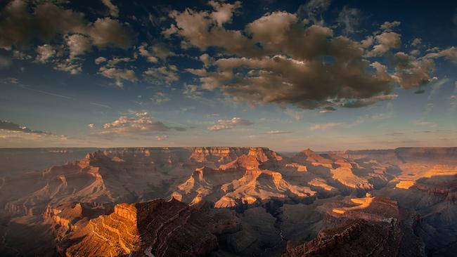 Die riesige Schlucht von weit oben, im Abendlicht werfen die Gesteinsformationen lange Schatten. Einige Wolken hängen am bläulich-violetten Himmel.