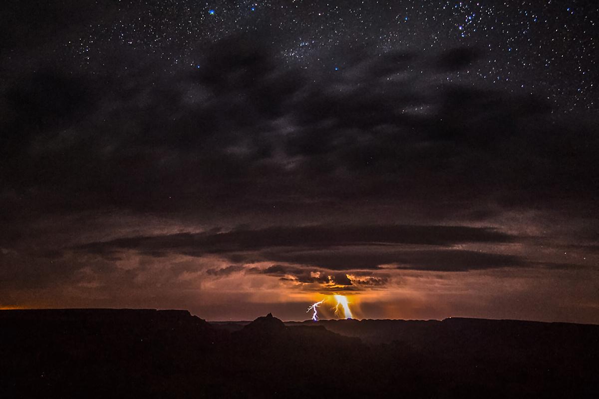 Oberhalb der schwarzen Silhouette des Canyons entlädt sich ein Blitz, der Wolken und Himmel orange erleuchtet. Über den dicken Gewitterwolken sind unzählige Sterne am Nachthimmel erkennbar.