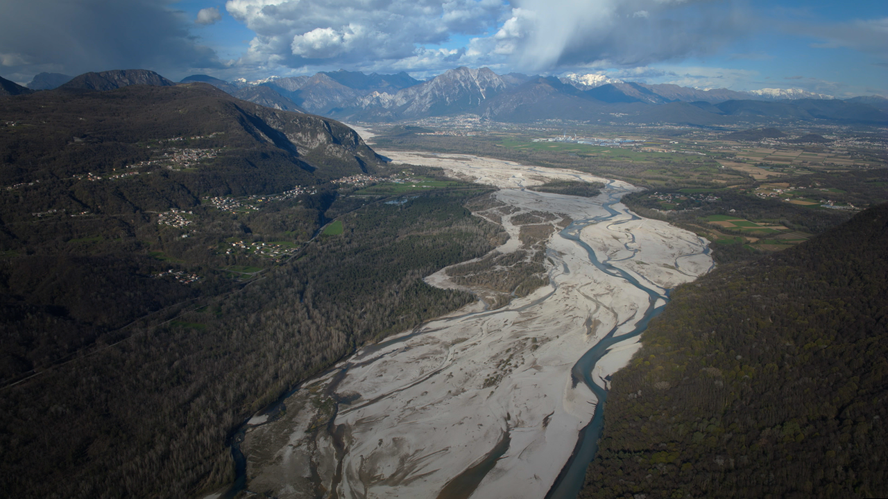 Von hoch oben: Ein riesiges Schotterbett mit verzweigten Gerinnen, das Wasser ist türkis. Im Hintergrund befinden sich hohe Berge.