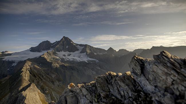 Ein schroffer Bergkamm führt zur markanten Pyramide des Großglockners. Teilweise liegt Schnee, zarte Wolkenschleier hängen am blauen Himmel.