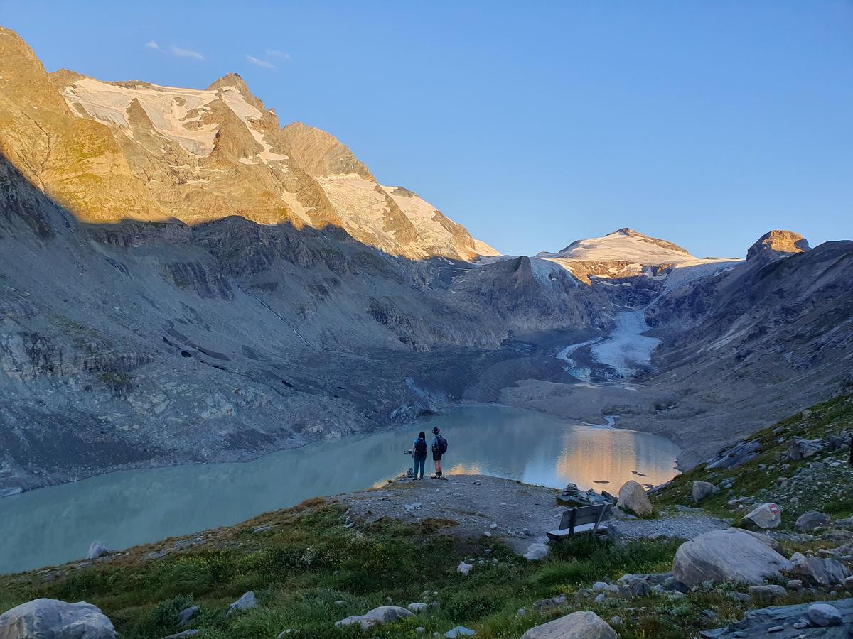 Zwei Menschen stehen am Ufer eines Schmelzwassersees in einem Talkessel, die Steilwände der Berge sind schroff. Am gegenüberliegenden Ufer des Sees liegt die Pasterze.
